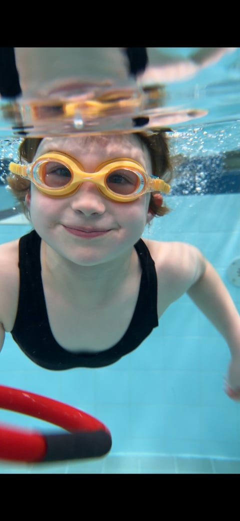 Child underwater in pool wearing yellow swimming goggles, smiling at camera while holding a red flotation device