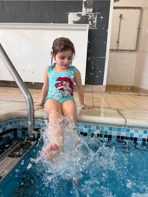 Young child in turquoise swimsuit splashing into an indoor swimming pool with metal railing visible