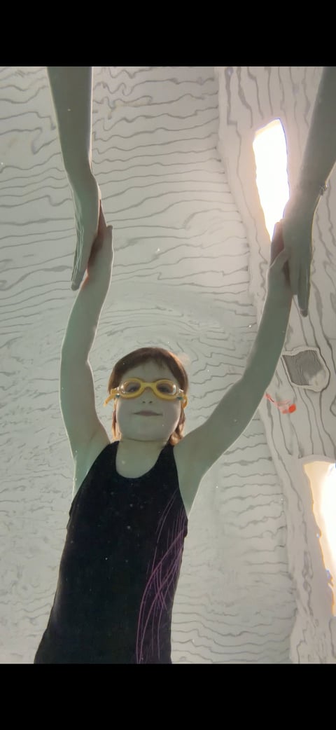 Child wearing yellow goggles with arms raised upward inside a narrow white textured space with bright light streaming in from above