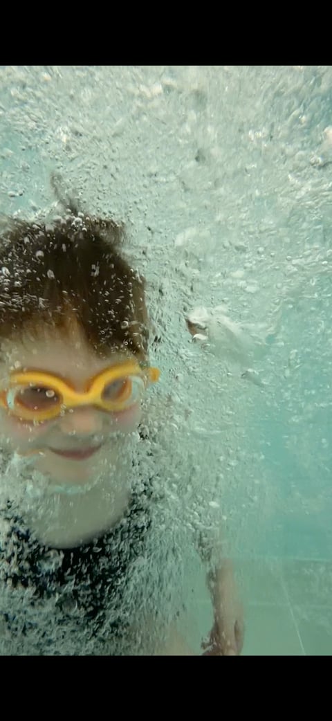 Child wearing yellow swim goggles underwater, surrounded by bubbles and clear turquoise water