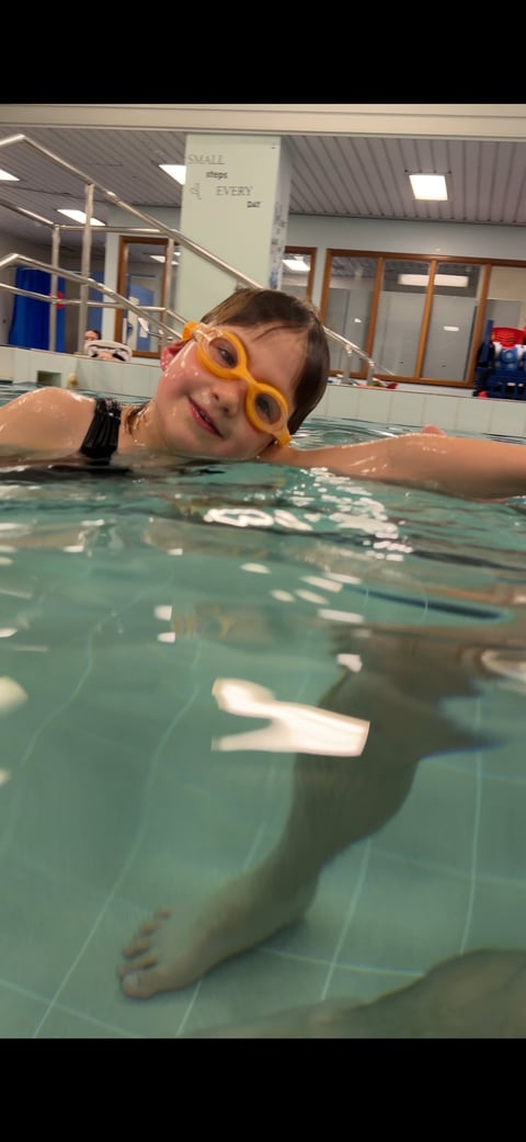Child in yellow swim goggles floating and smiling in indoor pool during swimming lesson