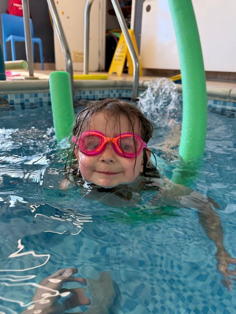 Young child in pink goggles swimming in pool with green foam noodles, smiling at camera