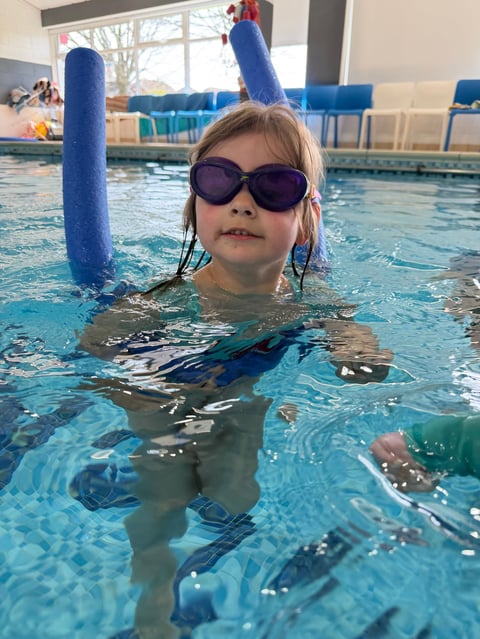 Child in swimming pool wearing sunglasses and holding blue flotation noodles during swim lesson at indoor pool facility