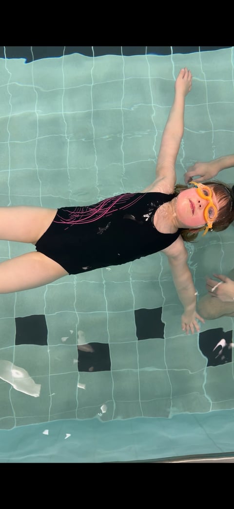 Child floating in a swimming pool wearing a black swimsuit and orange swim goggles, viewed from above
